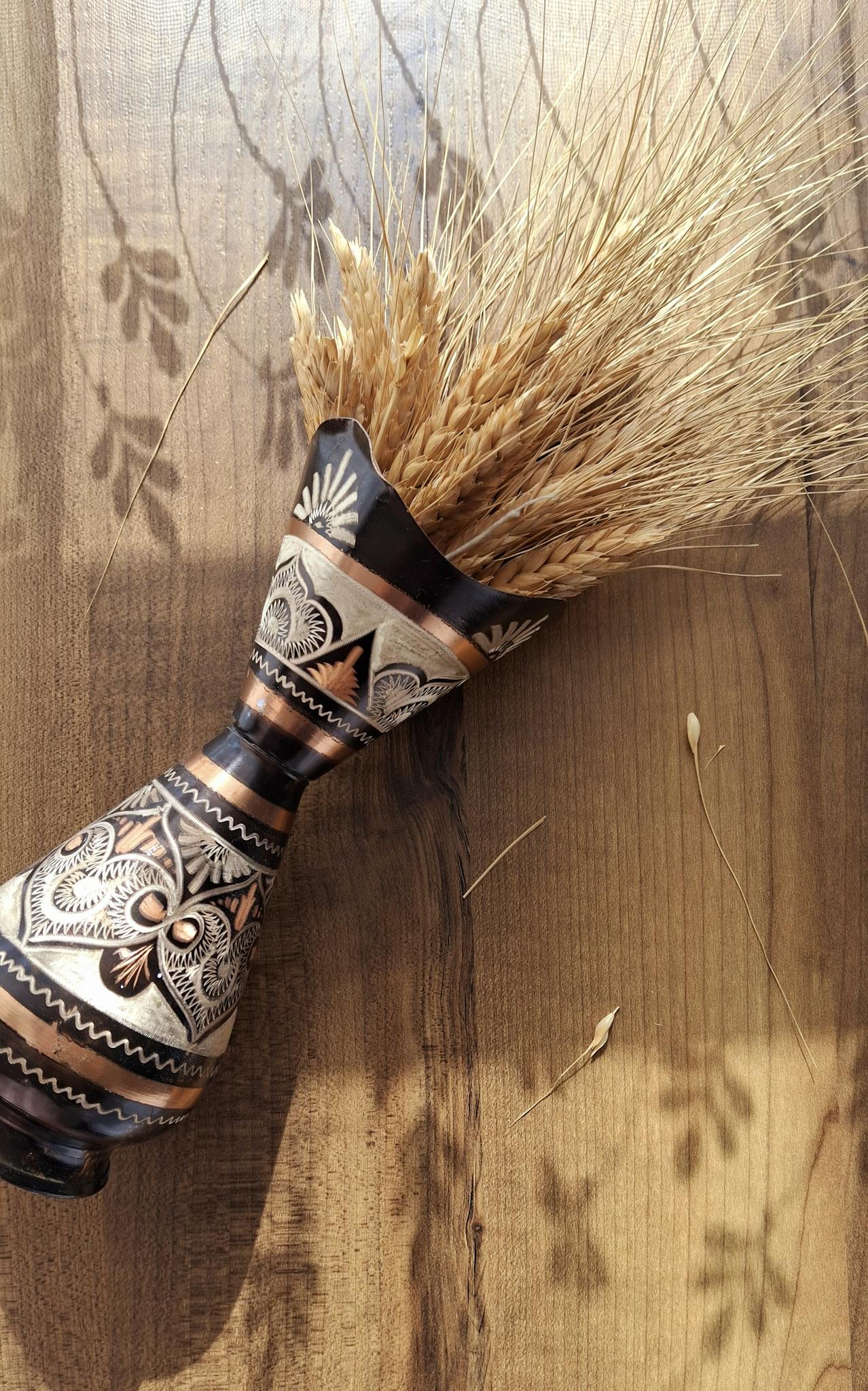 Decorative carved wooden vase with dried wheat on a wooden surface in natural light.